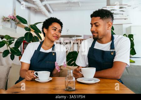 Ritratto del personale dell'Happy Cafe con un uomo ispanico e una barista di donna nera accanto ad un banco di panetteria. Immagini di ospitalità calde e diversificate, ideali per bar, panetteria o marketing di ristoranti. Mette in evidenza il lavoro di squadra, il servizio e l'accoglienza dei clienti. Foto Stock