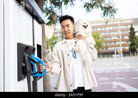 Uomo asiatico che trasporta il suo cane mentre usa l'ugello in una stazione di servizio in città. Un ambiente caldo e di tutti i giorni adatto per la proprietà di animali domestici e contenuti di vita urbana. Ideale per campagne sulla proprietà responsabile degli animali domestici e sulla vita familiare. Foto Stock