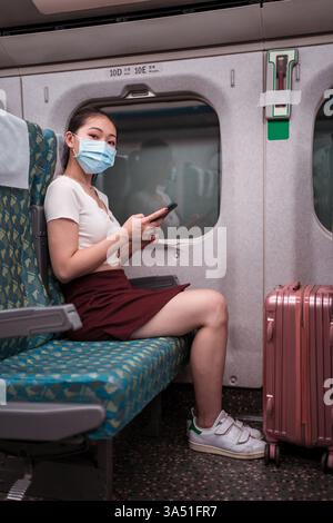 Donna asiatica che indossa una maschera di protezione viaggia in treno, utilizzando uno smartphone. L'immagine riflette la sicurezza degli spostamenti e i viaggi moderni durante i viaggi quotidiani. Adatto per articoli e campagne su viaggi, trasporti pubblici e precauzioni sanitarie. Foto Stock