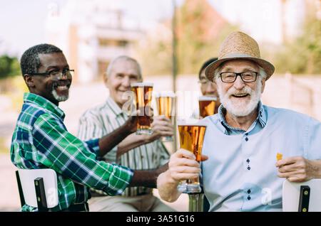 L'uomo nero sorridente con gli occhiali da sole siede con un gruppo di amici più grandi in un parco soleggiato. Stile di vita informale all'aperto con un'atmosfera multigenerazionale e socievole. Ideale per campagne di lifestyle, social e community. Foto Stock