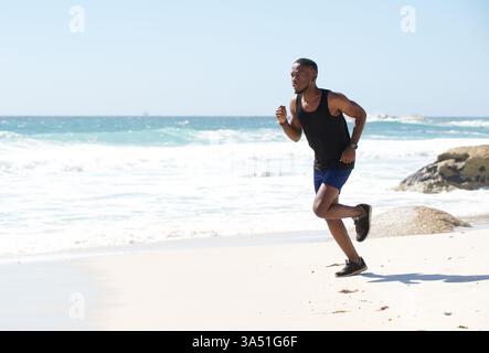 Sano afroamericano che corre lungo la spiaggia in riva all'acqua, catturando un vivace momento di fitness e di vita all'aperto. Adatto per campagne sportive, di benessere e motivazionali. Ideale per gli allenamenti in spiaggia e la grafica attiva. Foto Stock