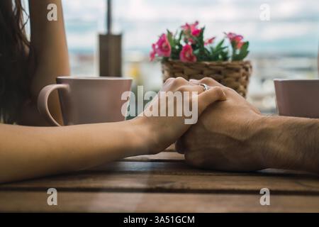 Una felice coppia ispanica tiene le mani sedute a un tavolo sul balcone di casa. L'intimo momento domestico trasmette calore e compagnia in un ambiente luminoso e rilassato. Foto Stock