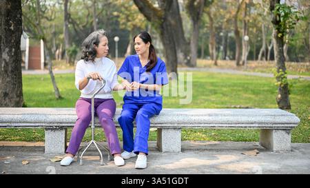 Infermiera asiatica in uniforme blu che tiene per mano una paziente asiatica anziana mentre si siede sulla panchina con il bastone quadruplo nel parco durante il giorno Foto Stock