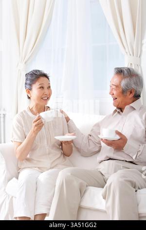 Una felice coppia cinese può gustare il tè insieme su un divano in un luminoso soggiorno. Ideale per lo stile di vita domestico, il relax e i momenti in famiglia. Foto Stock
