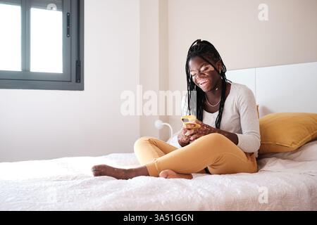 Positiva donna afroamericana in pantaloni gialli e maglione bianco seduta su un comodo letto vicino alla finestra mentre si utilizza lo smartphone chattando sui social Foto Stock
