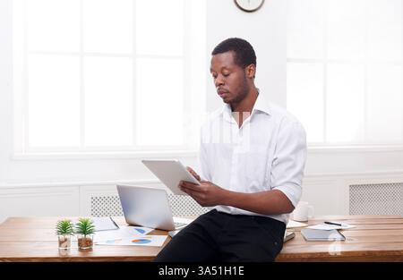 Uomo d'affari afroamericano concentrato in un ufficio moderno, che esplora le idee su un tablet con spazio di copia. La scena comunica leadership, innovazione e stili di lavoro digitali per contesti aziendali e tecnologici. Adatto per campagne di business, strategia e stile di vita professionale. Foto Stock