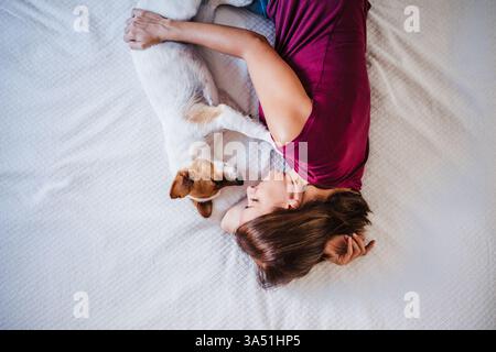 Allegra donna ispanica che gioca con un cane Jack Russell sul letto di casa. Un caldo momento domestico che mostra compagnia di animali domestici e spazio di vita rilassato. Perfetto per le campagne per lo stile di vita, la famiglia o la cura degli animali domestici. Foto Stock