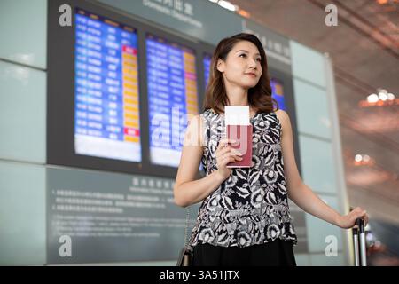 Donna cinese sorridente, in possesso di un passaporto e in piedi con i bagagli all'aeroporto. Questo ritratto pronto per il viaggio si adatta ai viaggi d'affari, ai viaggi internazionali e ai contenuti delle compagnie aeree. Foto Stock