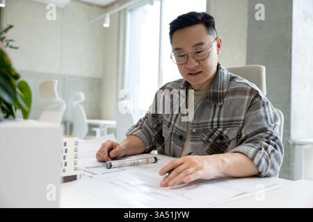 Un uomo asiatico esamina un modello di casa stampato in 3D in un ufficio luminoso e moderno in un laboratorio creativo. L'immagine sottolinea la progettazione architettonica, la tecnologia di stampa 3D e la collaborazione professionale intorno a un modello. Ideale per funzioni di innovazione delle costruzioni, sviluppo di progetti e lavoro di squadra ingegneristico. Foto Stock