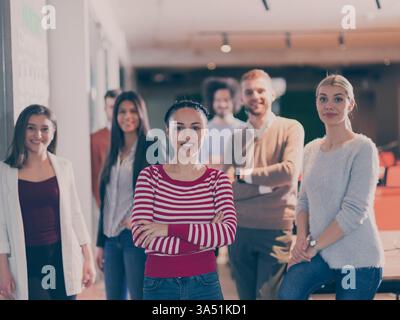 Ritratto di una bella donna d'affari che indossa un hijab in un ufficio moderno. Sorride alla telecamera mentre i colleghi multirazziali appaiono sullo sfondo, illustrando diversità e lavoro di squadra. Foto Stock