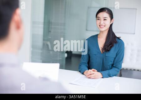 Una donna cinese sorridente si siede di fronte a un responsabile delle risorse umane asiatiche durante un colloquio di lavoro in un ufficio. La scena mette in evidenza il reclutamento, i colloqui e la comunicazione professionale. Foto Stock