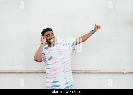 Un uomo afroamericano felice in una maglietta casual si erge su una strada contro una parete bianca, indossando le cuffie e ascoltando una canzone. Adatto per lo Street style, lo stile di vita urbano e campagne a tema musicale. Atmosfera vivace e conveniente per la tecnologia consumer e l'intrattenimento. Foto Stock