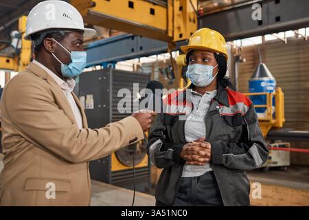 Ingegnere maschio nero che indossa un casco di sicurezza e una maschera per il viso tiene un microfono mentre intervista una lavoratrice donna nera in un magazzino. Il colloquio in loco evoca la trasmissione o la comunicazione aziendale in un ambiente industriale. Ideale per media, produzione e storytelling sul luogo di lavoro. Foto Stock