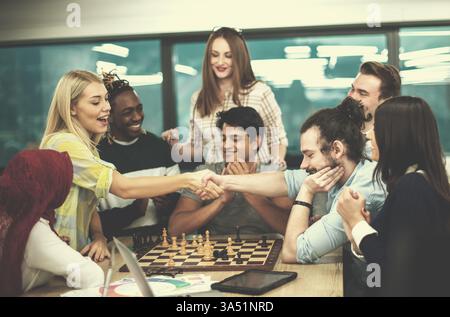Scena d'ufficio variegata con un uomo d'affari misto e una donna caucasica che stringono la mano dopo una pausa scacchi. Il momento sottolinea il lavoro di squadra, la strategia e la diversità all'interno di un ufficio moderno. Ideale per la cultura delle startup, la leadership e la grafica di collaborazione aziendale. Foto Stock