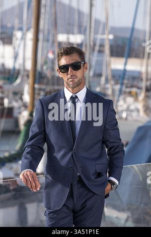 Il fiducioso uomo d'affari ispanico con gli occhiali da sole si trova accanto a una ringhiera di vetro in un porto soleggiato. Vestito con una tuta acuta, trasuda leadership professionale mentre si appoggia all'aperto contro la ringhiera. Questa elegante immagine executive si adatta allo stile di vita aziendale, alle impostazioni del porto e alle campagne di ritratti all'aperto. Foto Stock