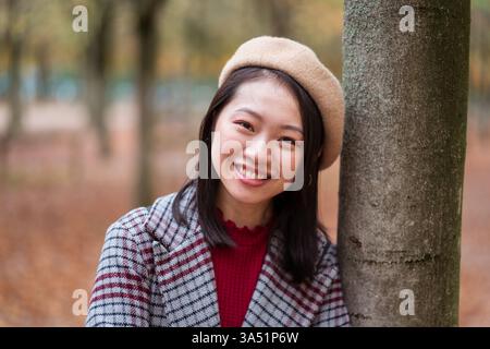 Donna asiatica sorridente in un berretto e cappotto a scacchi si appoggia su un albero in un parco autunnale. Questo ritratto stagionale è perfetto per la moda, lo stile di vita e il marketing stagionale. Ideale per l'uso editoriale o commerciale, con un look sicuro ed elegante per esterni. Foto Stock