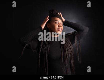 Donna nera sorridente in piedi con un panino intrecciato sullo sfondo scuro di uno studio. Presenta un look elegante e contemporaneo adatto per ritratti e fotografia di moda. Questo ritratto trasmette fiducia e bellezza moderna. Foto Stock