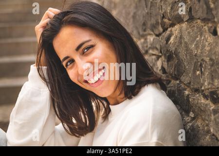Primo piano ritratto di una giovane donna sorridente seduta sui gradini. La posa rilassata e l'espressione amichevole lo rendono ideale per lo stile di vita, la bellezza e i momenti quotidiani in un ambiente urbano. Foto Stock