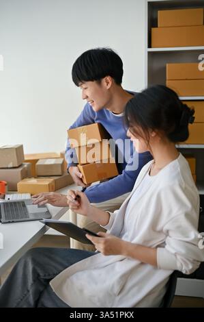 Felice coppia thailandese lavora da un ufficio di casa, controllando e confermando gli ordini online su un portatile. Un'immagine pratica per l'e-commerce, le piccole imprese e i contenuti di avvio, con la collaborazione di coppia a casa. Foto Stock
