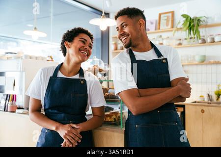 Calda scena da caffe' con un uomo ispanico sorridente e una donna nera che serve pasticceria e caffe'. Il duo di baristi al bancone crea un ritratto di ospitalità invitante e diversificato per le campagne di ristorazione. Foto Stock
