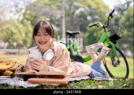 Donna asiatica sorridente che indossa le cuffie intorno al collo utilizzando lo smartphone sdraiato sul tappetino da picnic vicino alla bicicletta nel parco durante la giornata di sole Foto Stock