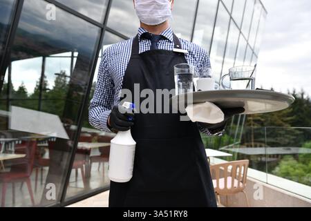 Cameriere che pulisce un tavolo del ristorante con spray disinfettante, indossando una maschera e guanti. La scena riflette l'igiene e la sicurezza nell'ospitalità e nella nuova normalità. Ideale per campagne di sicurezza per la ristorazione, pulizia del ristorante e protocolli sanitari. Foto Stock