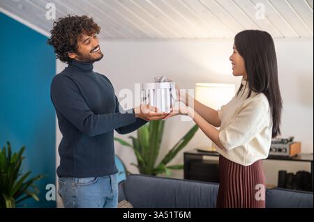 Un gioioso momento di vacanza a casa come un uomo asiatico regala un regalo a una donna ispanica sorridente. La scena è all'interno con una luce calda, che enfatizza lo scambio di regali e le festività. Un'immagine versatile per Natale, occasioni familiari e relazioni diverse. Foto Stock