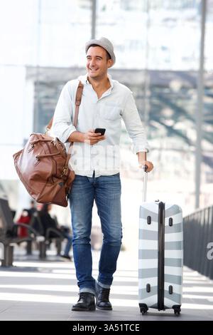 L'allegro ispanico in un terminal dell'aeroporto cammina con una borsa e un bagaglio mentre si guarda via. Ha in mano uno smartphone, catturando un momento di viaggio sincero in un ambiente di vita frenetico. Ideale per campagne sui viaggi moderni e sull'utilizzo dello smartphone durante il trasporto. Foto Stock