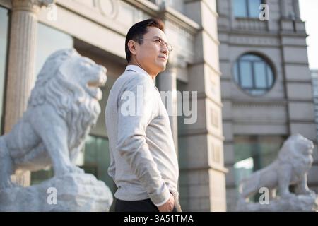 Uomo d'affari cinese di mezza età con una mano in tasca si erge accanto a una statua di leone fuori da un edificio in una strada della città durante il giorno. L'ambiente urbano all'aperto comunica leadership e fiducia per i temi aziendali e della vita cittadina. Un ritratto in vita adatto per il lavoro e la professionalità. Foto Stock
