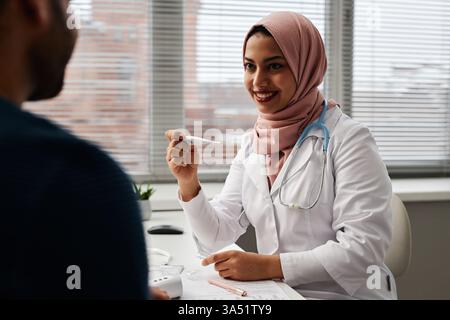 Giovane e allegro medico che indossa l'hijab e tiene in mano un termometro digitale mentre guarda un paziente in una clinica. La scena trasmette cure compassionevoli e servizio medico professionale. Perfetto per comunicazioni sanitarie, cliniche e donne in medicina. Foto Stock