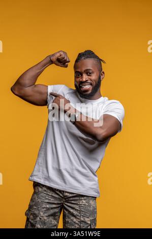 Un uomo afroamericano sorridente sta in piedi e punta al bicipite, mostrando la forza della parte superiore del corpo. Il ritratto dell'atletica mette in evidenza il fitness e il benessere, ideale per la palestra, lo sport o le campagne di benessere. Ideale per la promozione e la pubblicità attiva sullo stile di vita. Foto Stock