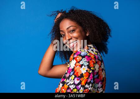 Vista laterale di una allegra donna afroamericana con abiti eleganti che toccano i capelli ricci e guardano la fotocamera con un sorriso scherzoso. Moda vivace e ritratti di stile, ideali per campagne allegre e sicure di sé. Foto Stock