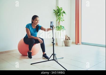 Una donna ispanica sorridente si siede su una palla da ginnastica registrando un video a casa. Perfetto per fitness, social media e contenuti influencer. Ideale per il benessere, gli allenamenti domestici e i materiali di marketing per lo stile di vita. Foto Stock
