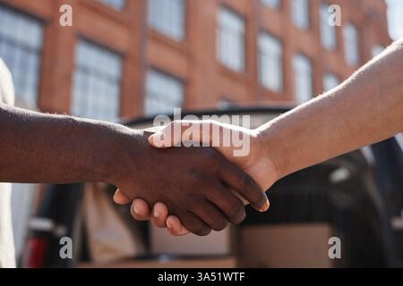 Vista laterale primo piano di una stretta di mano tra diversi professionisti all'aperto con un furgone per consegne sullo sfondo. La scena comunica partnership e fiducia per le campagne aziendali, logistiche ed e-commerce. Foto Stock