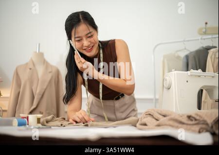 Una donna asiatica, professionale e impegnata, stilista di moda o sarta sta parlando al telefono con il suo cliente mentre lavora sul suo computer portatile nel suo atelier Foto Stock