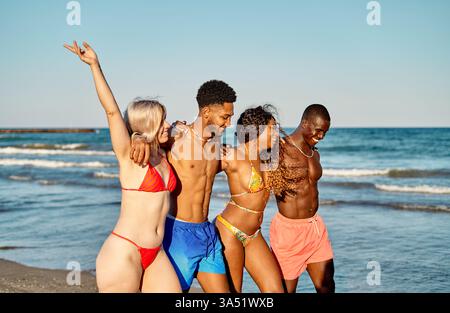 Un gruppo di amici diversi in una passeggiata sulla spiaggia soleggiata, braccia sollevate in un momento celebrativo. Condividono una spensierata giornata estiva in riva al mare, incarnando l'amicizia e le vibrazioni di viaggio. Perfetto per lo stile di vita, i viaggi e il tempo libero all'aperto. Foto Stock