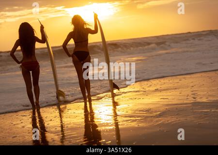 Due surfiste, asiatiche e caucasiche, si stagliano con tavole da surf su una spiaggia baciata dal sole al tramonto. Guardano lontano, creando una scena serena e aspirazionale perfetta per lo stile di vita in spiaggia o per le campagne sportive. Ideale per l'estate, i viaggi e gli sport oceanici con un'atmosfera tropicale. Foto Stock