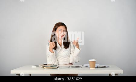 Una donna d'affari asiatica sorridente siede alla sua scrivania e regala un pollice, isolata su uno sfondo bianco. Questa immagine in stile ritratto offre sicurezza e risultati eccellenti in ambienti professionali. Adatto per le comunicazioni aziendali, il successo sul luogo di lavoro e il branding positivo. Foto Stock