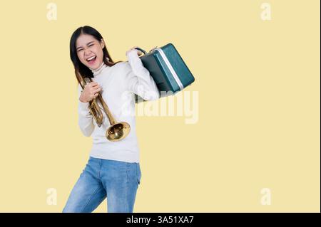 Il allegro trombettista asiatico si erge su uno sfondo giallo brillante in studio, reggendo una tromba. Questo vivace ritratto si adatta alla musica, al branding degli strumenti, ai concerti e ai materiali didattici. Foto Stock