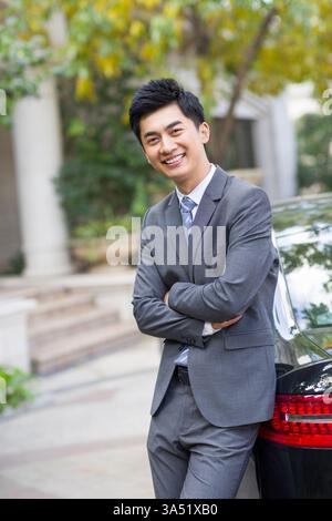 Uomo d'affari cinese con le braccia incrociate si appoggia contro un'auto in un garage. Questa posizione sicura suggerisce prontezza e mobilità professionale. Foto Stock