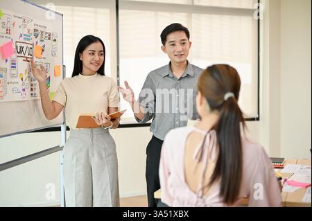 Sorridendo gli sviluppatori di tecnologie asiatiche maschili e femminili in piedi e presentando il loro nuovo progetto durante la riunione del team in ufficio Foto Stock