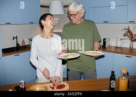 Coppia di anziani che preparano allegramente un pasto insieme nella cucina moderna, condividendo un momento felice e sorridendo calorosamente. La cucina include ingredienti freschi e utensili Foto Stock