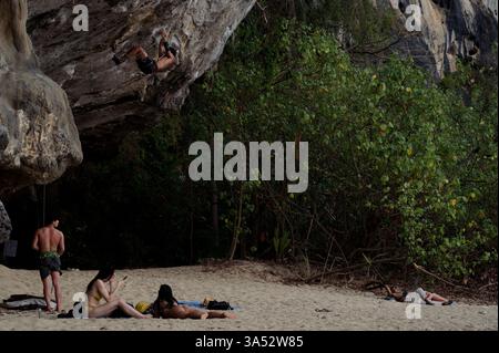 L'arrampicatore sale su una ripida sporgenza, mentre gli altri si rilassano sulla spiaggia sabbiosa sotto la scogliera coperta dalla giungla Foto Stock
