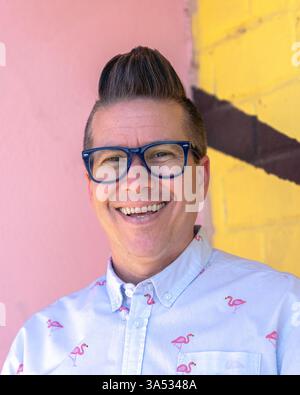 Ritratto ravvicinato di una persona sorridente con capelli corti marroni, occhiali con cornice blu e camicia button-up azzurra con fenicottero rosa Foto Stock