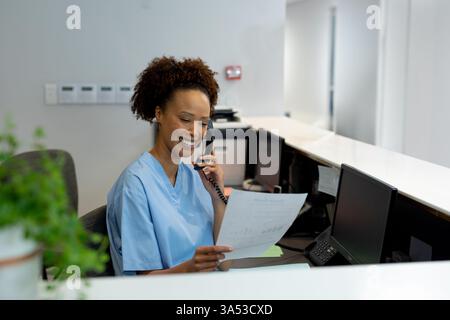 Il receptionist dell'ospedale in scrub sorride mentre parla al telefono e tiene un documento Foto Stock