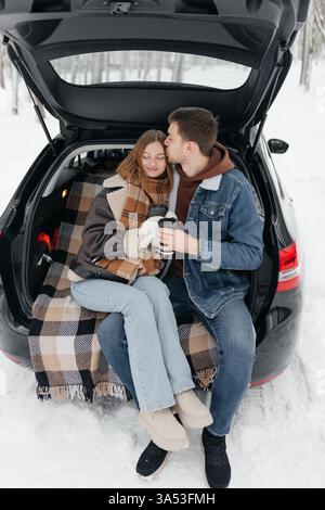 In un pittoresco paese delle meraviglie invernali, una coppia condivide con piacere bevande calde e dolci coccole nel bagagliaio della propria auto, creando un'atmosfera davvero accogliente Foto Stock
