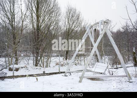 Vecchio altalena in legno ricoperta di neve nelle fredde giornate invernali con alberi senza fronzoli e recinzione sullo sfondo. Concetto di luoghi abbandonati, cambiamento stagionale, e. Foto Stock