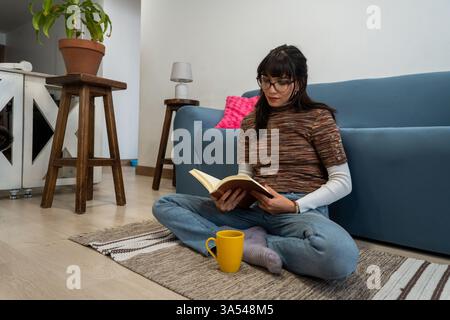 Giovane donna seduta sul pavimento a leggere un libro con una tazza di caffè accanto a lei, godendosi un tranquillo pomeriggio a casa Foto Stock