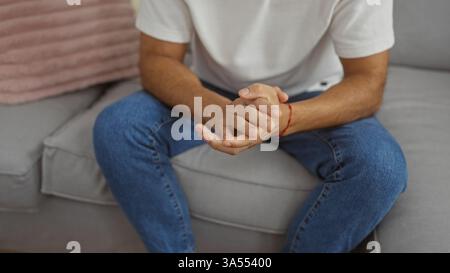 Giovane ispanico seduto in casa con le mani strette, vestito in modo informale con jeans e camicia bianca, creando un'atmosfera rilassata e premurosa a casa Foto Stock