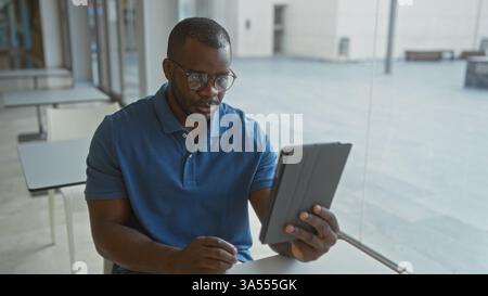 Giovane uomo dall'espressione seria che guarda un tablet in un ambiente di ufficio moderno, circondato da luce naturale e arredamento minimalista, si impegna in un ambiente di lavoro mirato Foto Stock